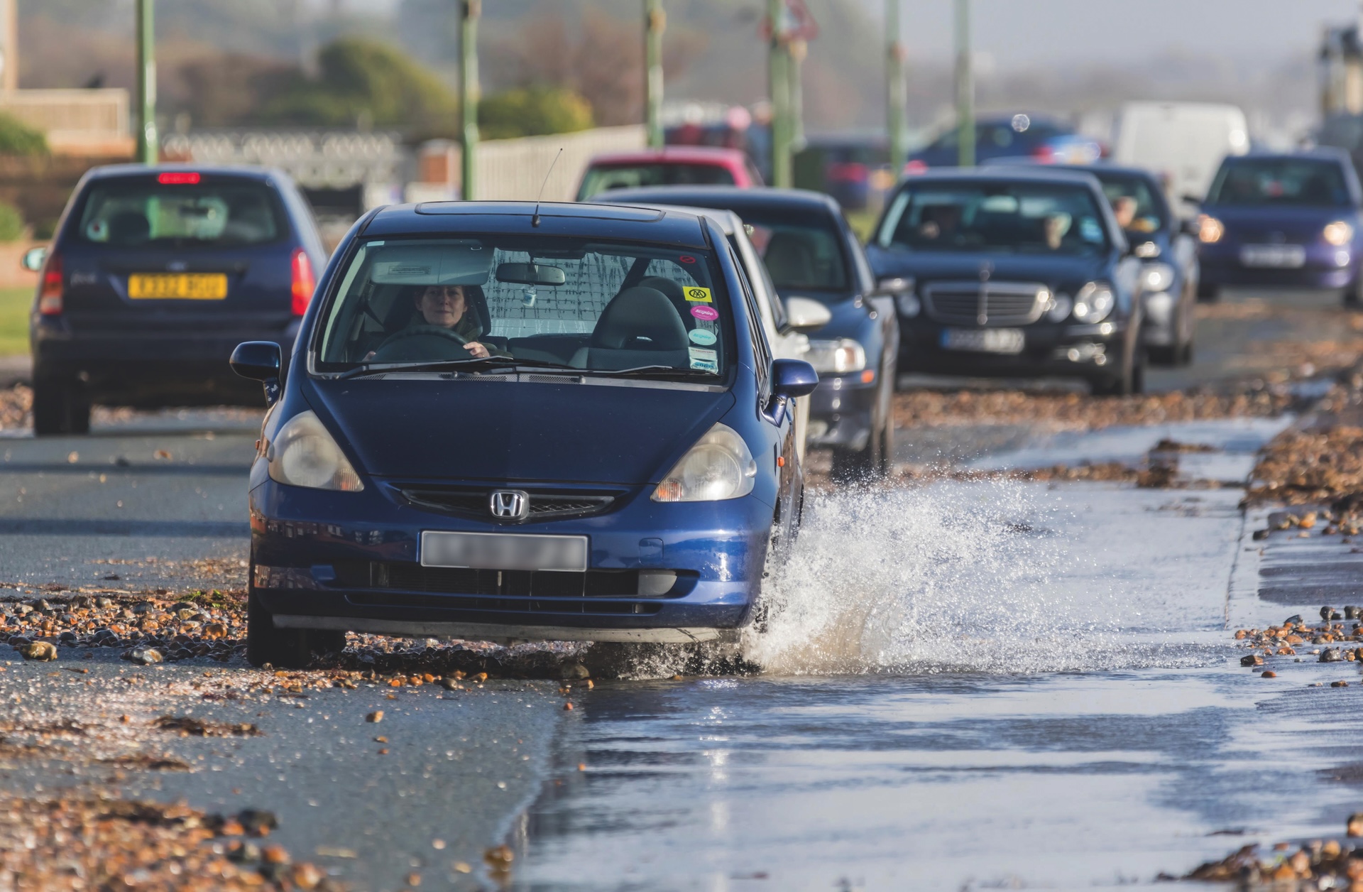 Car Driving Slowly Through Water On Flooded Road After Heavy Rain And High Tide Sea Water And Pebbles From The Beach Onto The Road In West Sussex, UK.